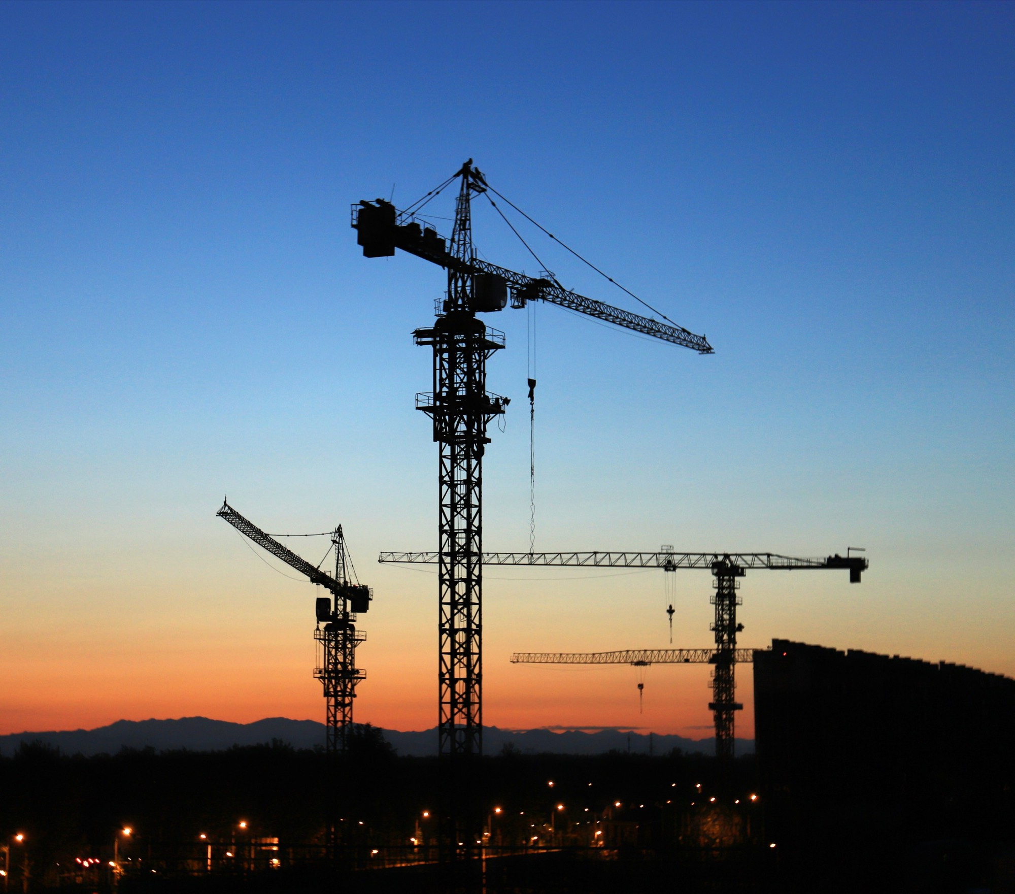 Tower cranes against a blue-hour sky fading to an orange horizon above a mountain ridge and city lights